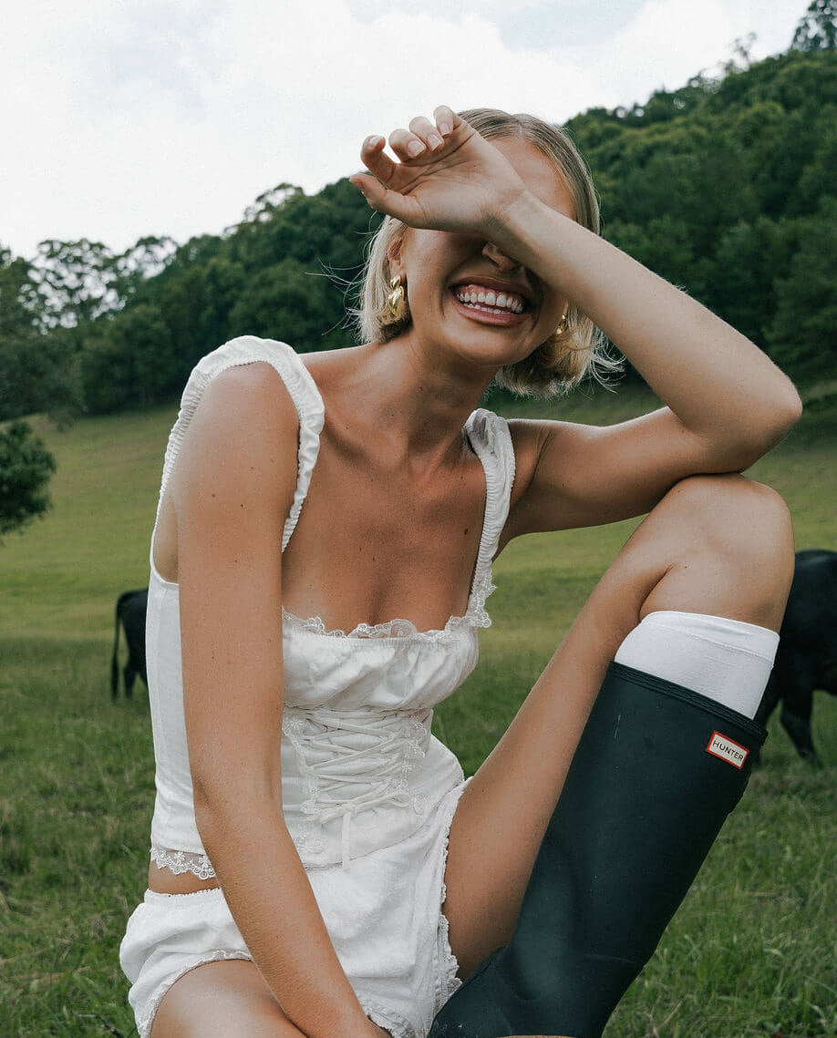 A woman sitting in a grass field surrounded by cows. She is laughing.