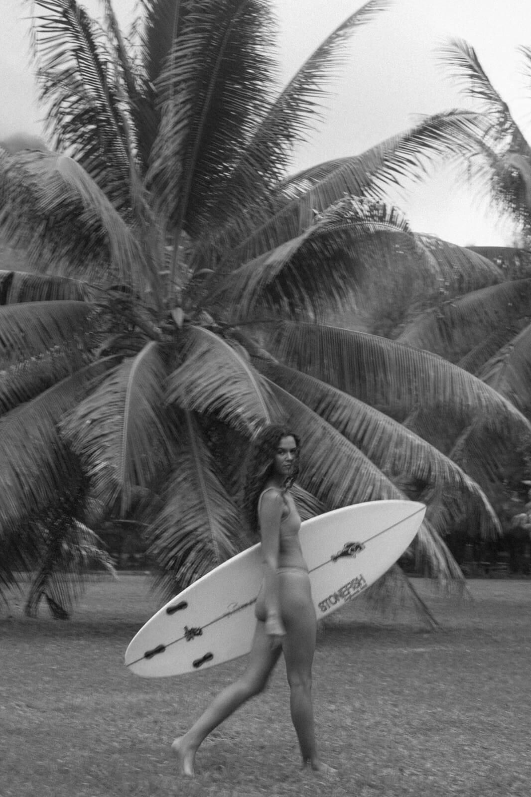 Woman in a bikini is walking past palm trees carrying a surfboard