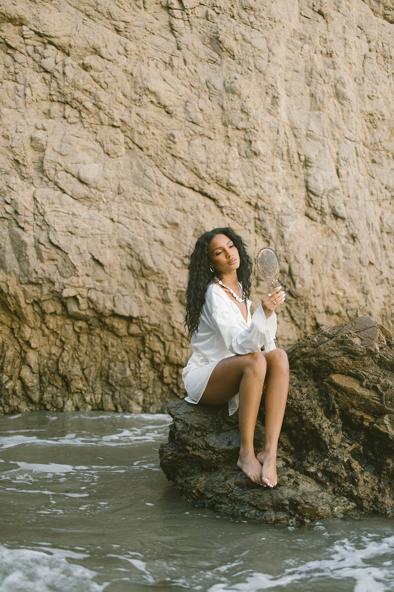 Woman in a white dress holding a mirror while she sits on a rock by the sea.